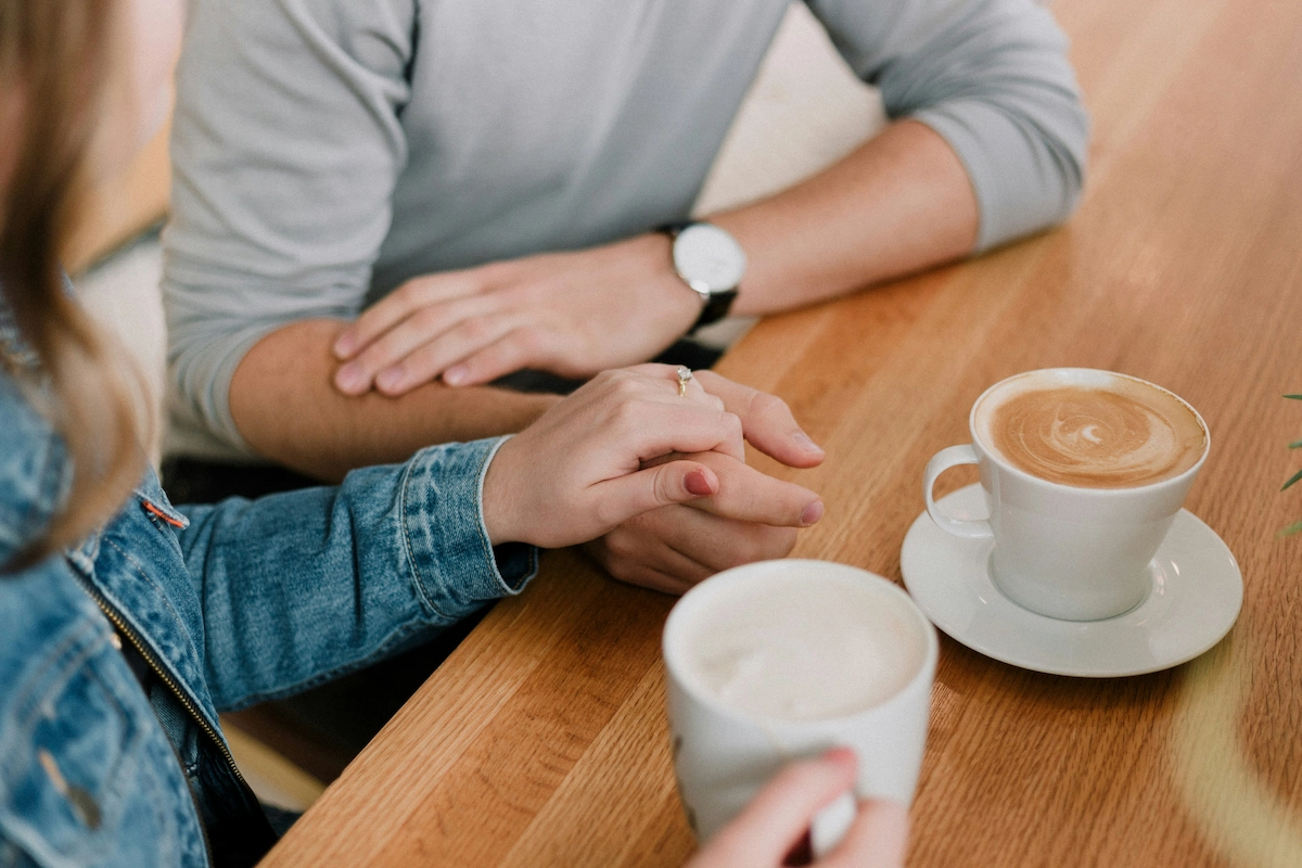 A photo of a couple sitting at a table over coffee, with their hands gently layered over each other's. Our featured image for our article on 'how to help a partner who gambles'