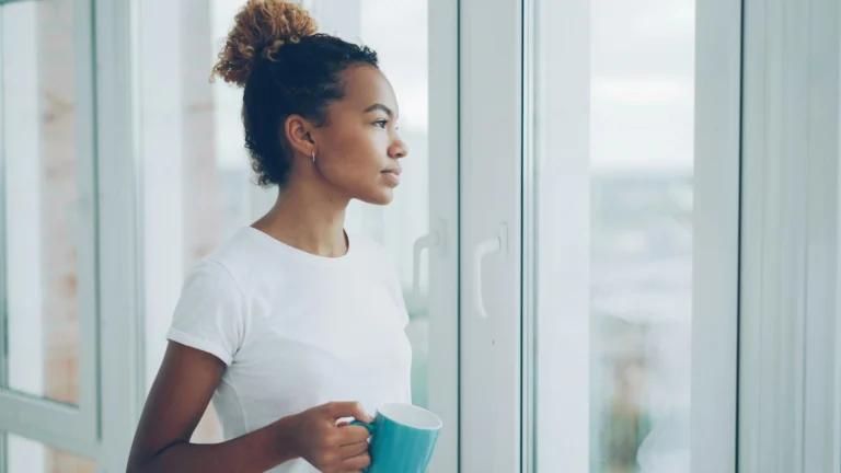 Photo of a woman standing with her cup of tea, distracted, staring out of the window. Our featured image for our article on 'recognising the signs of a gambling addiction'